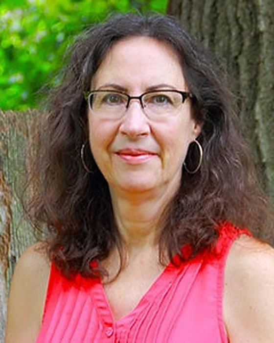 Tammy, the artist behind Rock River Stitches, smiling outdoors beside a tree